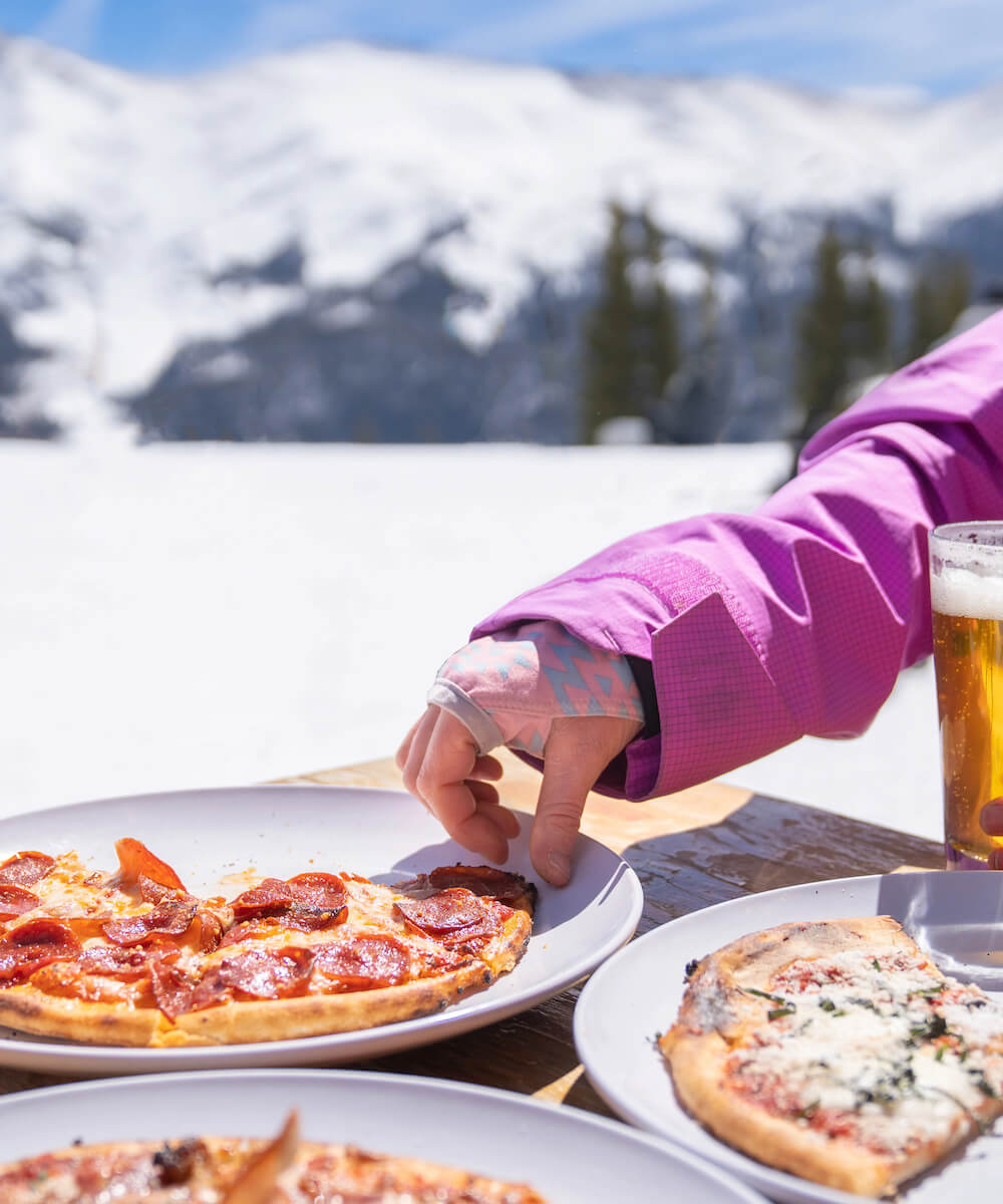 woman's hand reaching for pizza with a snowy mountain in the background